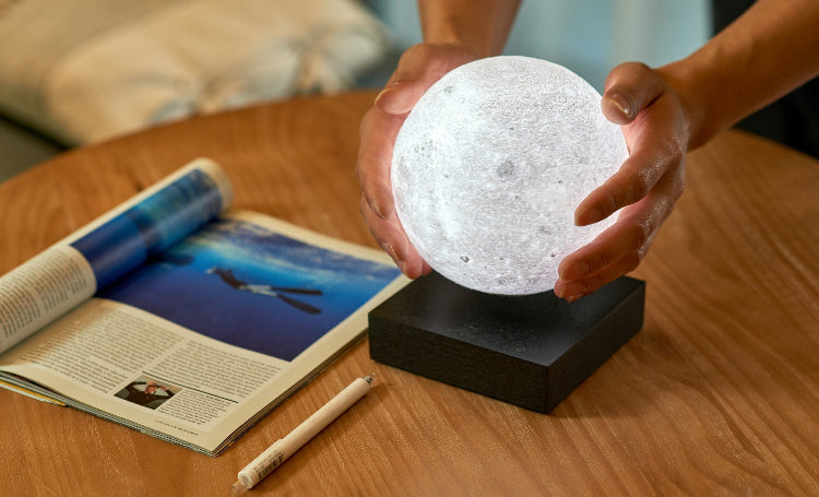 Person holding a moon-shaped lamp on a wooden table with an open book and pen.