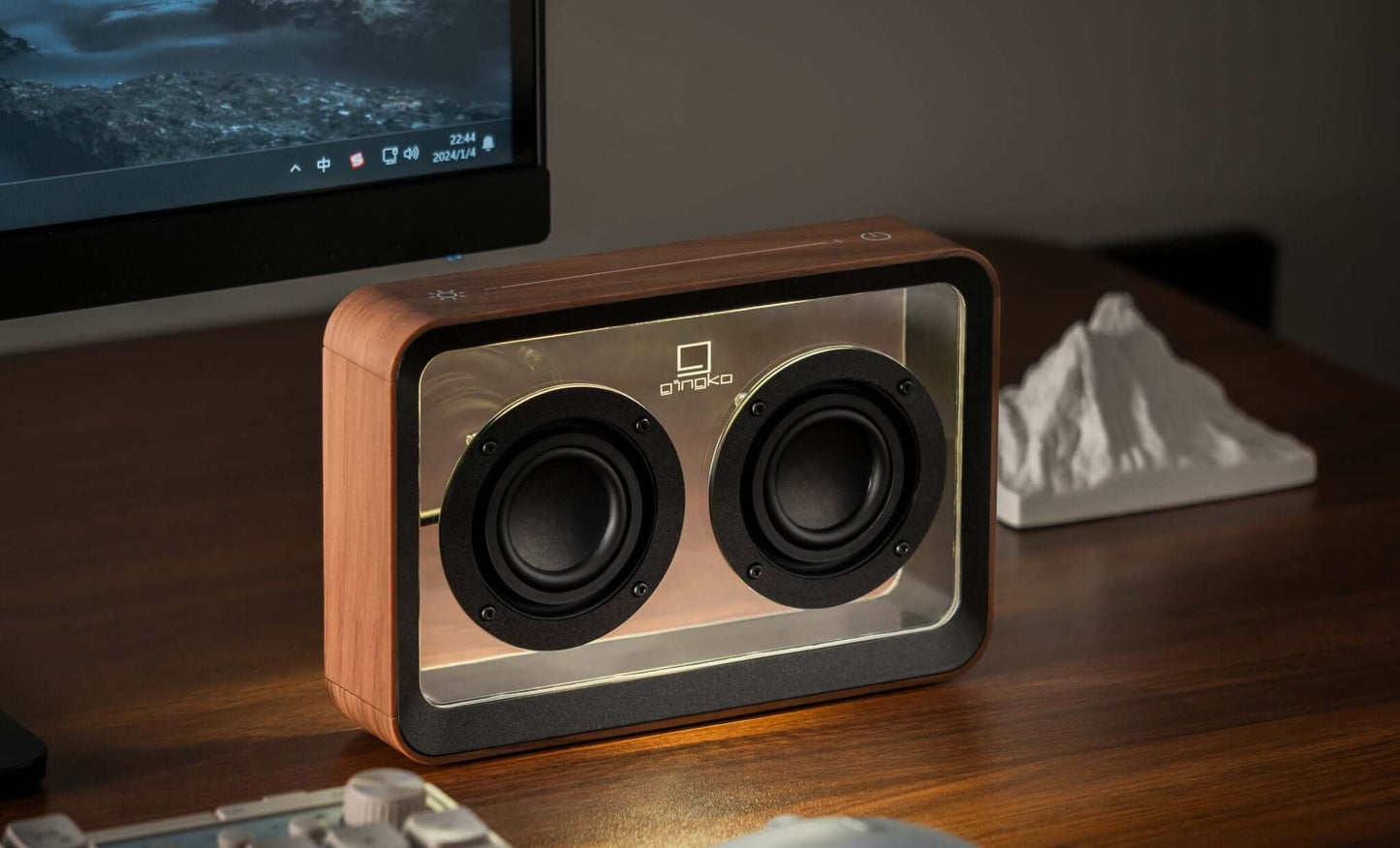 A transparent, acrylic Mage See-Through Bluetooth Speaker with a warm yellow light beneath the top cover, placed on a wooden surface next to a computer keyboard and monitor.