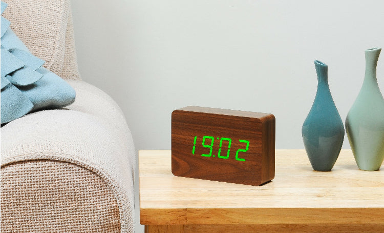 A sleek wooden alarm clock with an LED display showing the time '10:00' displayed on a wooden surface next to a blue vase.