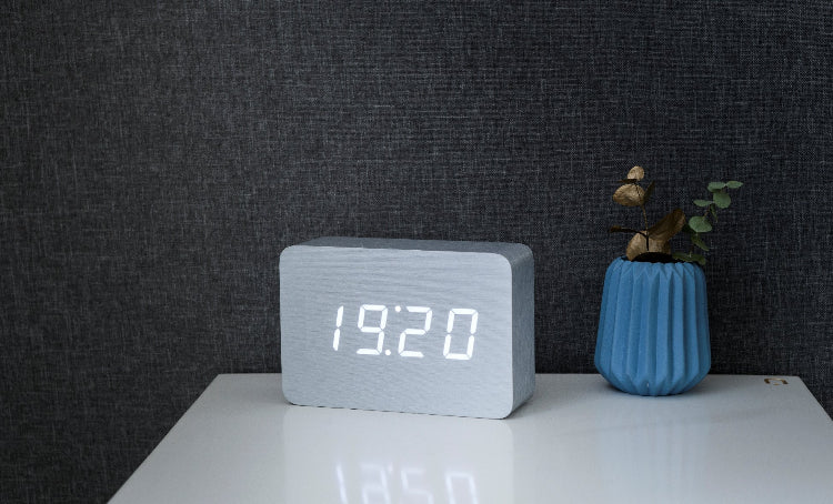 A sleek wooden alarm clock with an LED display showing the time '10:00' displayed on a wooden surface next to a blue vase.
