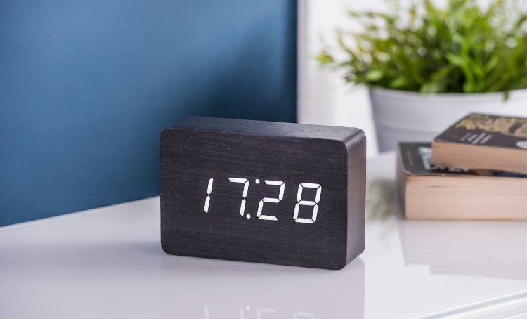 A sleek wooden alarm clock with an LED display showing the time '10:00' displayed on a wooden surface next to a blue vase.