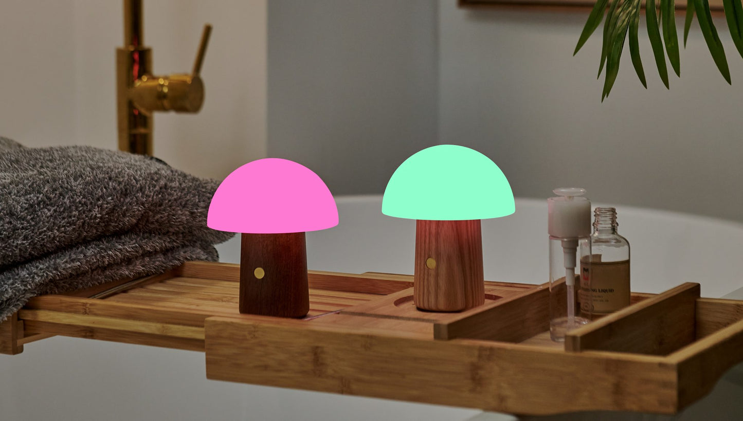 Two mushroom-shaped LED lamps with a walnut and white ash wood finish, one emitting a pink light and the other a green light, placed on a wooden tray on a table.