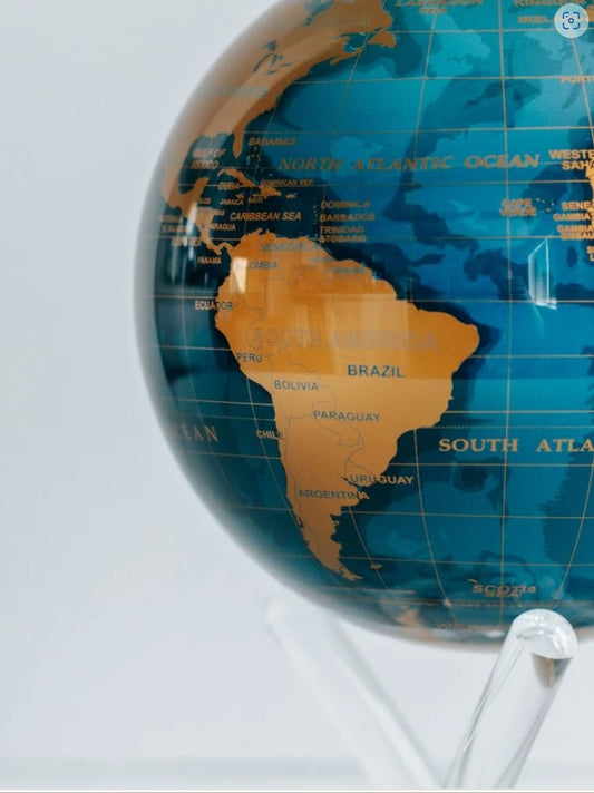 A contemporary rotating globe with cerulean blue oceans and gold metallic landmasses, powered by ambient light, displayed on a clear stand.