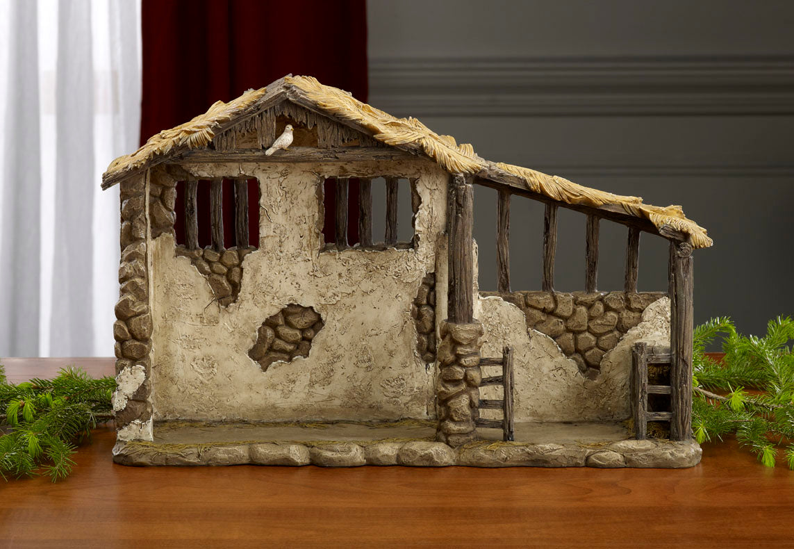 A detailed, hand-sculpted, lighted nativity stable with stone and stucco-style walls, wood beams, a thatched roof, and a hay-strewn floor.