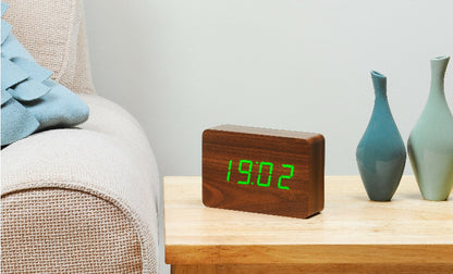 A sleek wooden alarm clock with an LED display showing the time '10:00' displayed on a wooden surface next to a blue vase.