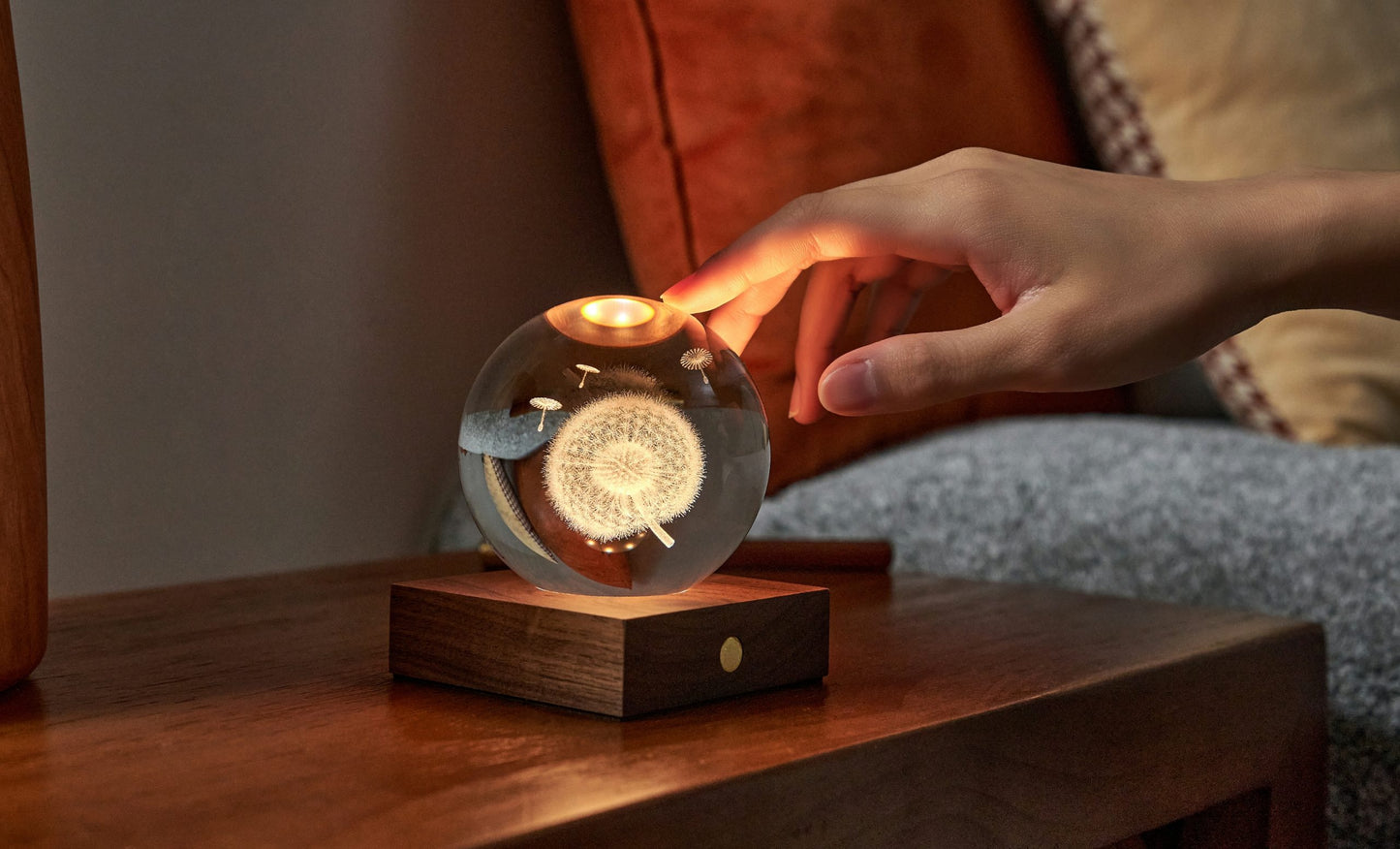 A hand touching a crystal ball lamp with a dandelion design, sitting on a wooden base.
