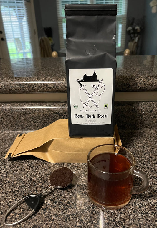 A bag of Kingdom Of Arms Coffee with a graphic of a coffee plant and coffee cup, alongside a clear cup of brewed coffee and a spoon on a kitchen countertop.
