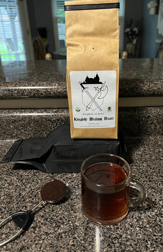 A bag of Kingdom Of Arms Coffee with a graphic of a coffee plant and coffee cup, alongside a clear cup of brewed coffee and a spoon on a kitchen countertop.