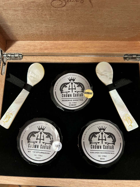 A luxury caviar gift box set open to reveal three tin containers of caviar and two mother-of-pearl spoons, all displayed on a black interior with a wood exterior.