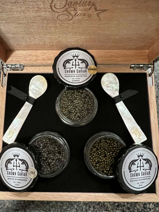 A luxury caviar gift box set open to reveal three tin containers of caviar and two mother-of-pearl spoons, all displayed on a black interior with a wood exterior.