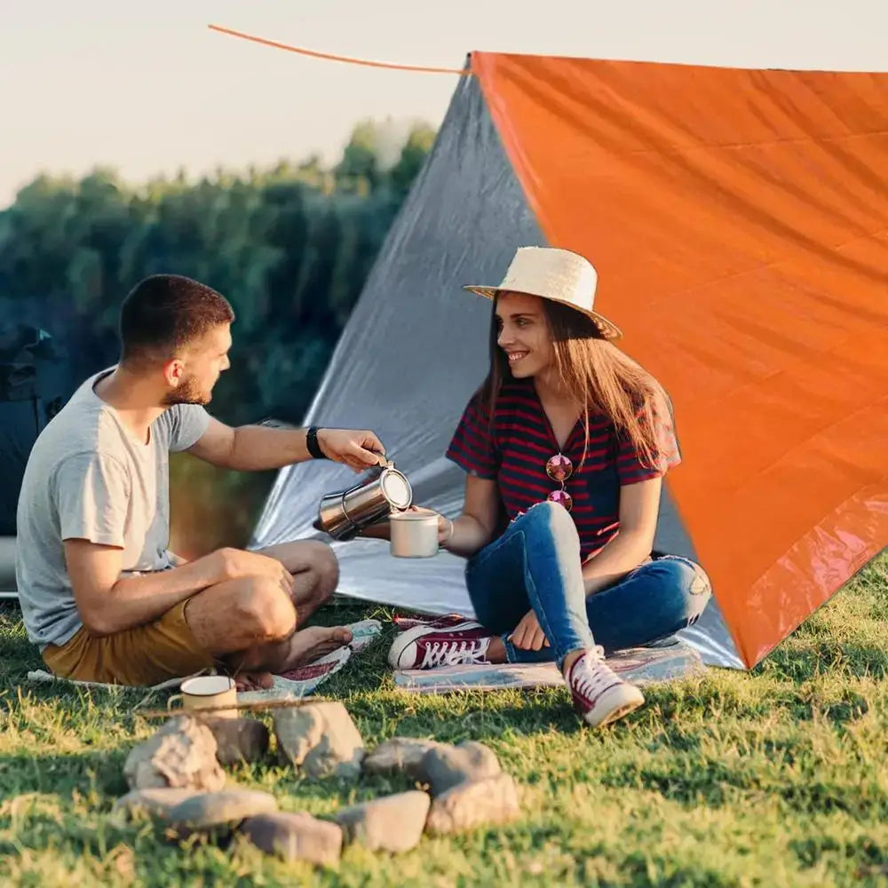 Couple enjoying coffee in front of outdoor emergency survival tent and blanket during camping trip.