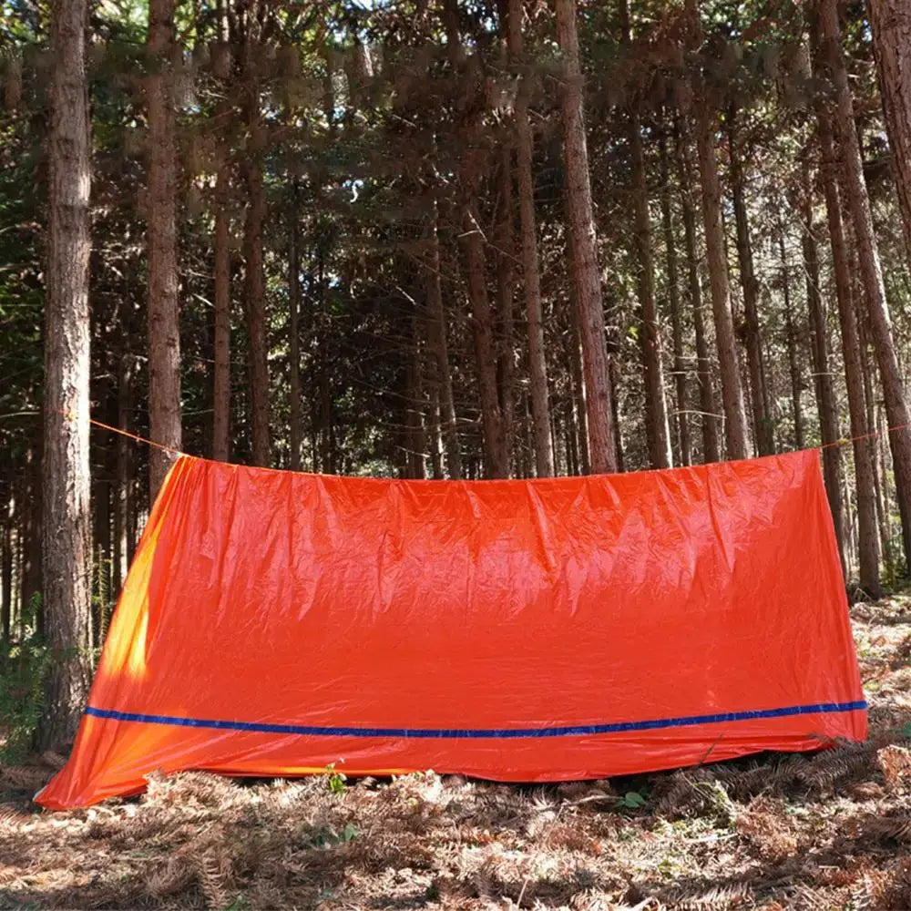 Outdoor Emergency Survival Tent and Blanket set up in a forest, showcasing its bright orange color for visibility.