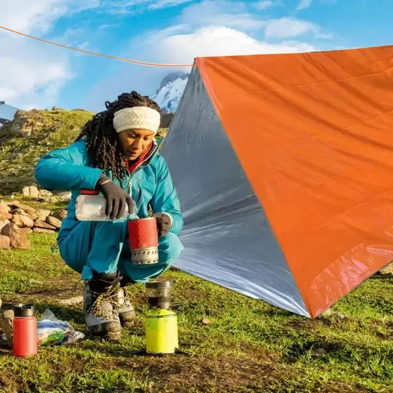 Outdoor Emergency Survival Tent and Blanket being used by a person setting up a camping stove in nature.