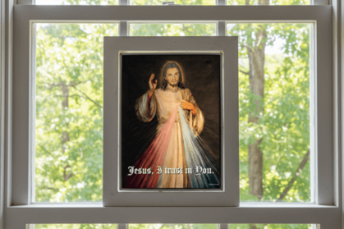 Framed artwork of a religious figure of 'Jesus, I trust in You' text, placed on a windowsill with a view of trees.