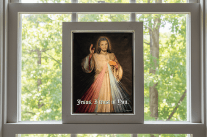 Framed artwork of a religious figure of 'Jesus, I trust in You' text, placed on a windowsill with a view of trees.