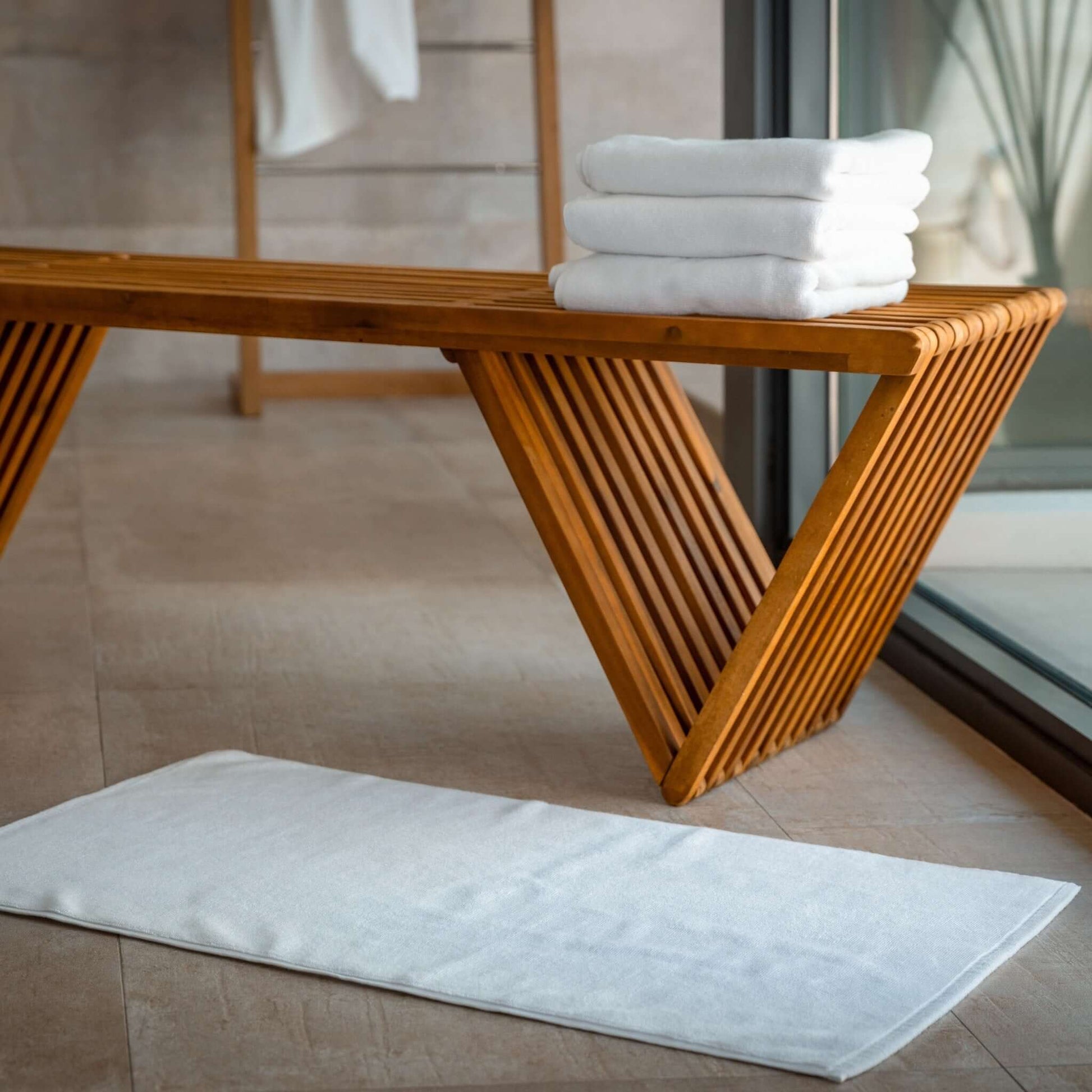 Luxury organic bath mats placed under a wooden bench with neatly stacked towels in a modern bathroom setting.