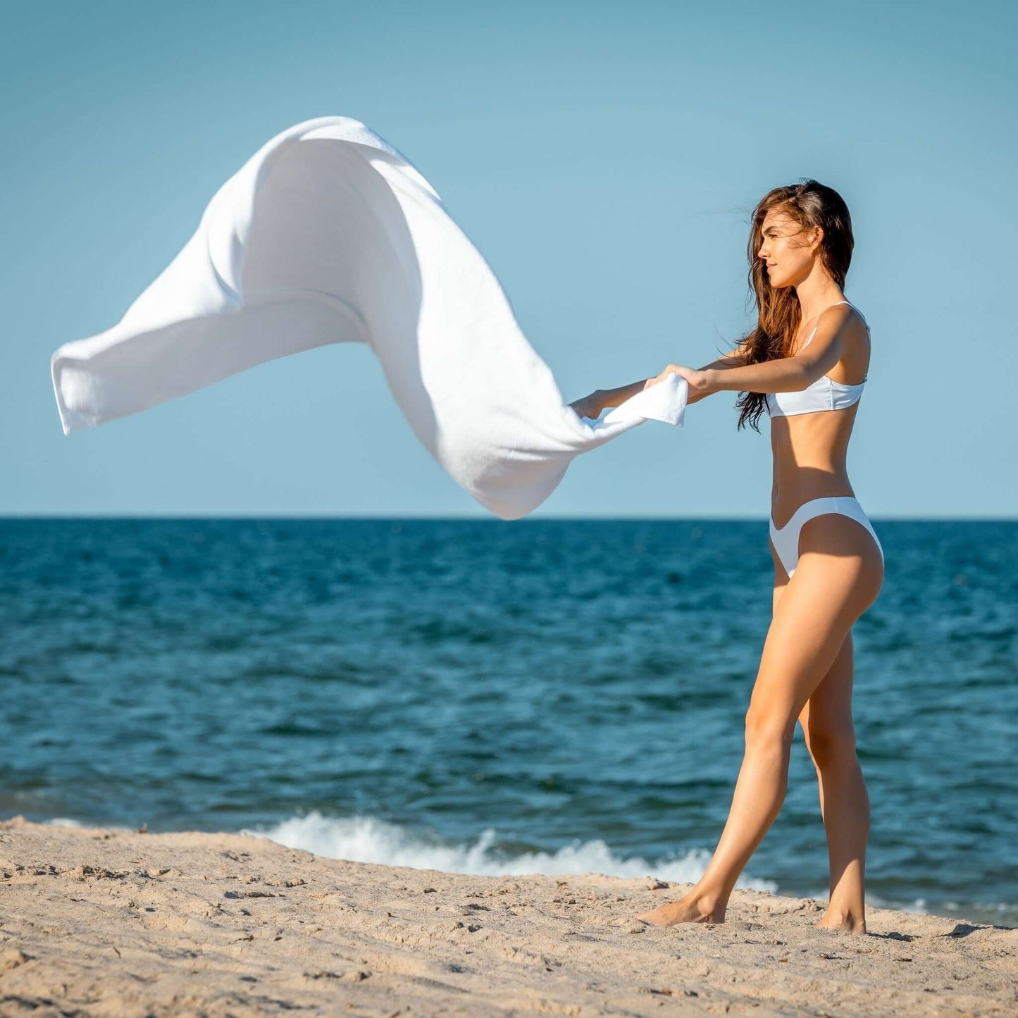 Woman at the beach enjoying the breeze while holding luxury organic bath sheets in white, capturing an eco-friendly lifestyle.